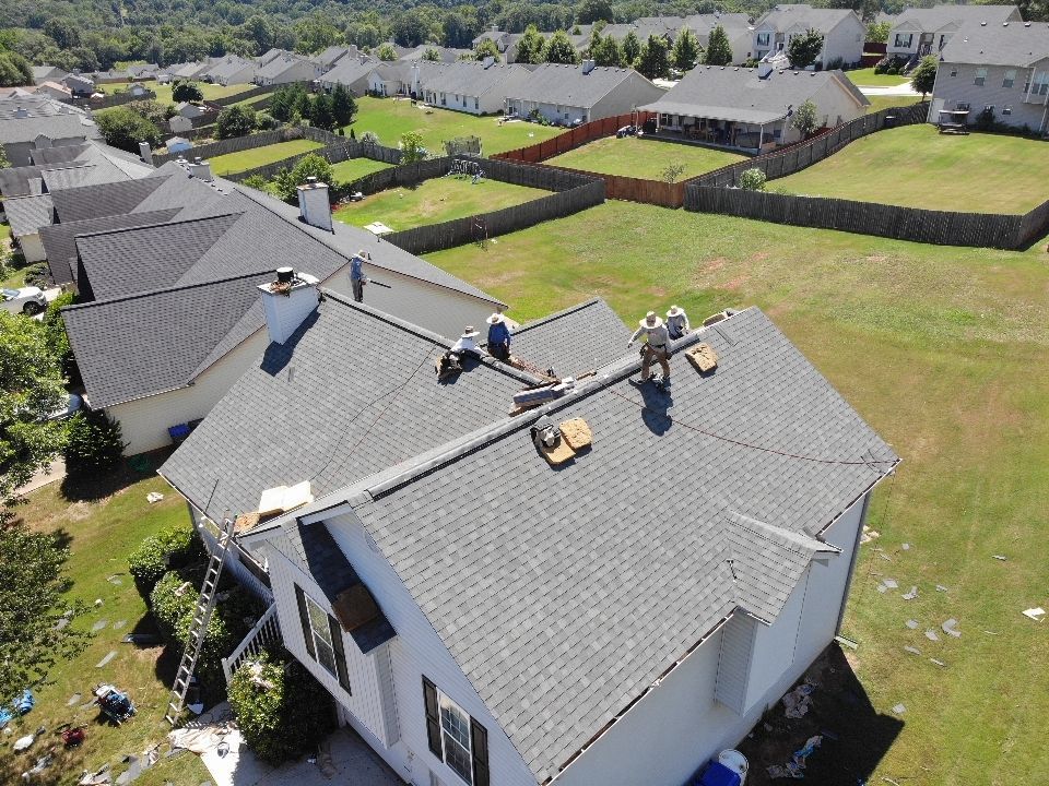 Roofers working on a gray shingled roof of a house. Other houses and green yards are in the background.