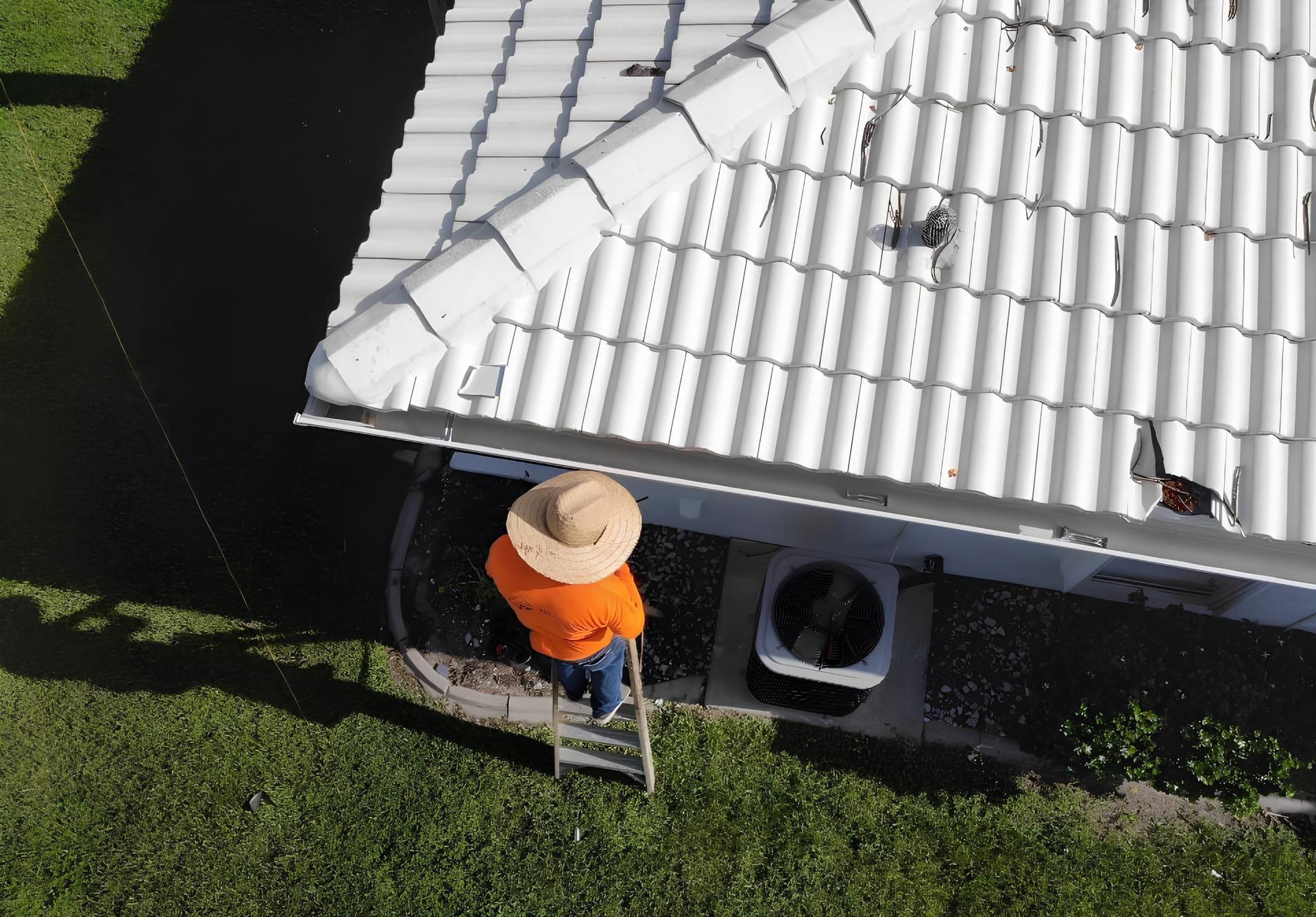 Person on a ladder near a white roof, wearing an orange shirt and straw hat, next to an air conditioning unit.