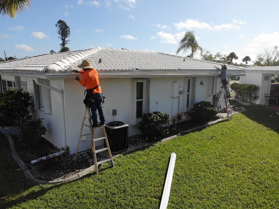 Two workers installing gutters on a white, single-story building with a white tile roof. One worker is on a ladder.