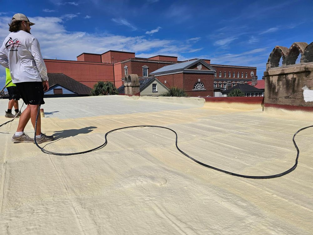 Two workers on a flat rooftop spraying insulation. Tan, textured roof with buildings and blue sky background.