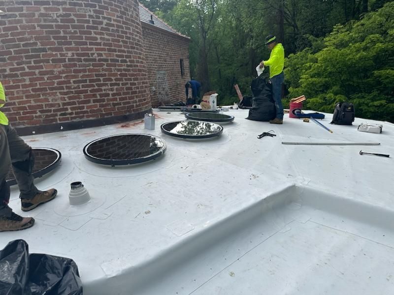 Workers on a white flat roof near a brick building. One wears a neon vest. Black garbage bags and tools are present.